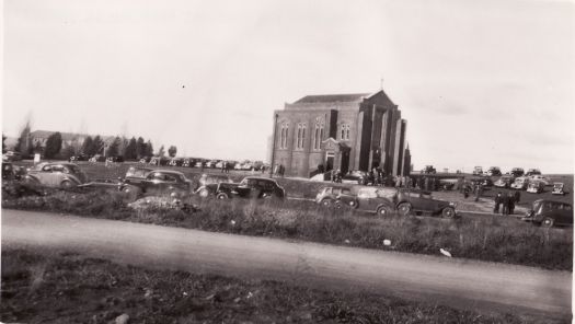 St Paul's Church of England, Manuka, standing in an open paddock. There are a number of cars parked near the church.