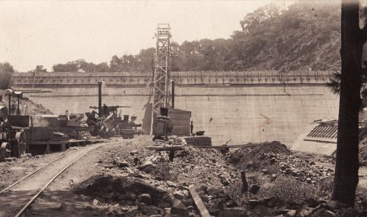 Shows the original Cotter Dam under construction and a rail line leading up to the site on the southern side of the Cotter River.