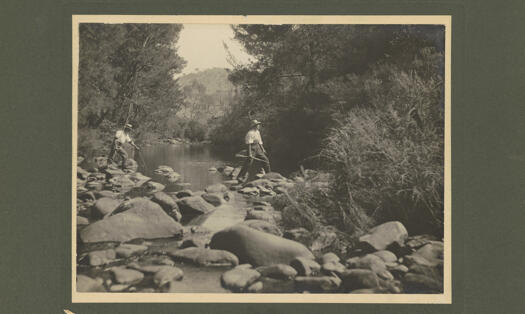 Two men crossing the Cotter River, stepping across stones. One is holding a gun.
