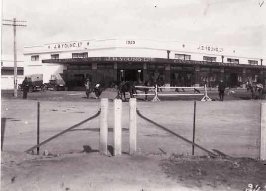 J.B. Young's on the corner of Giles Street and Jardine Street, Kingston. There are two horses attached to a hitching rail in front of the building and two early model cars parked in the street.