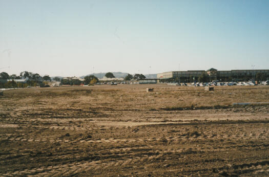 View from The Valley Way to Gungahlin Marketplace. A car park is visible in front of the shops and to its left. Houses along Anthony Rolfe Avenue are visible at rear. Gribble Street is at left as is Joint Services building.