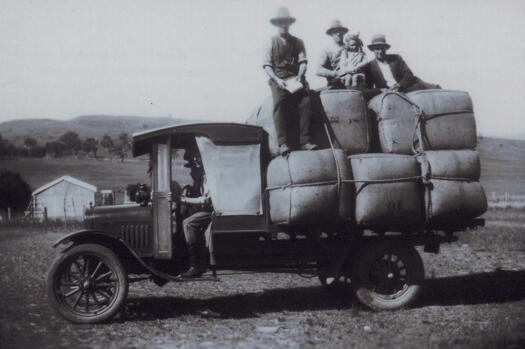 Oswald Dixon sitting in the front of a truck loaded with bales of wool from Kama. Three men and a girl are on top of the bales.