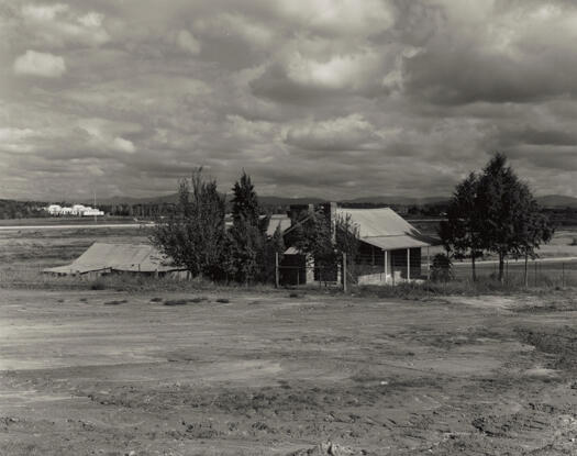 View of Blundells Cottage, undergoing restoration, before Lake Burley Griffin was formed. Parliament House is visible in the background.