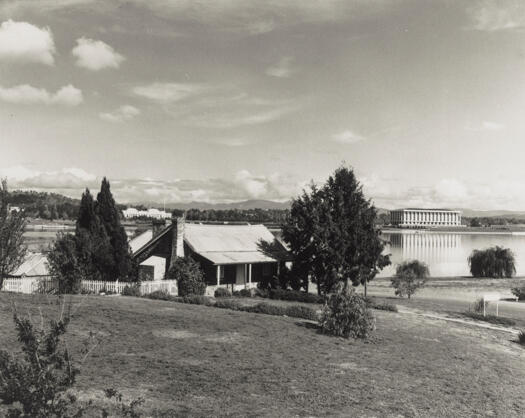 View of Blundells Cottage in April 1964, after restoration, Lake Burley Griffin has been formed and Parliament House is visible in the background.
