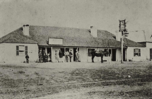 Single storey building. Several people standing outside. Horse tethered.