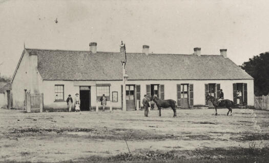 Single storey building with six external doors. People and horses outside.