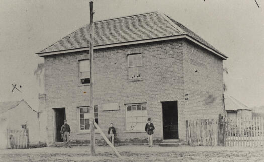 Two storey brick building built 1836. Three people standing outside.