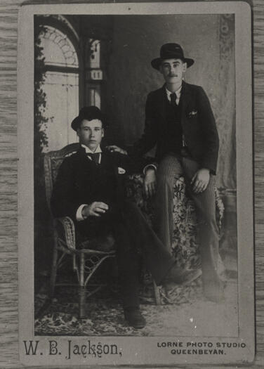 Studio portrait of two well-dressed young men. One sitting and one perched. Early 1890s.