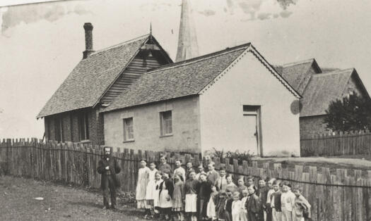 Students and teacher standing in front of fence. Original school and 1865 addition shown.