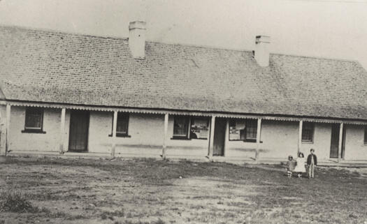 Single storey building with 3 doors and two chimneys. Three children standing outside.