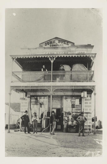 Front view of two storey building with top storey verandah and fancy ironwork trim. Four men standing outside and two men on top verandah.