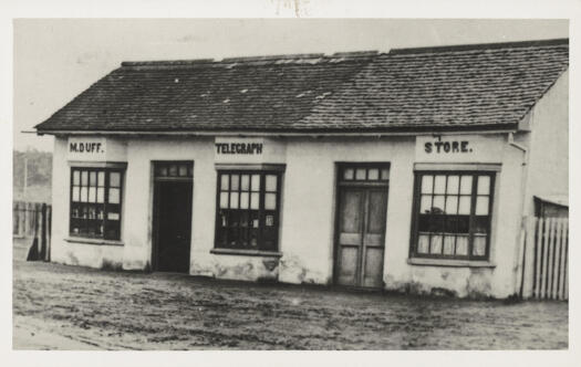 Single storey building with two doors and three bay windows. Sign reads 'M.J. Duff. Telegraph Store.'