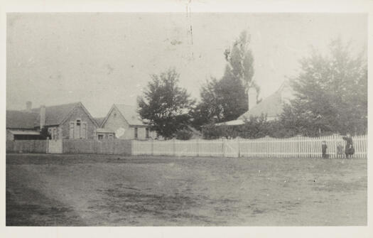 The school is on the far left and a fence runs along the street to a house with a more stylish white picket fence.